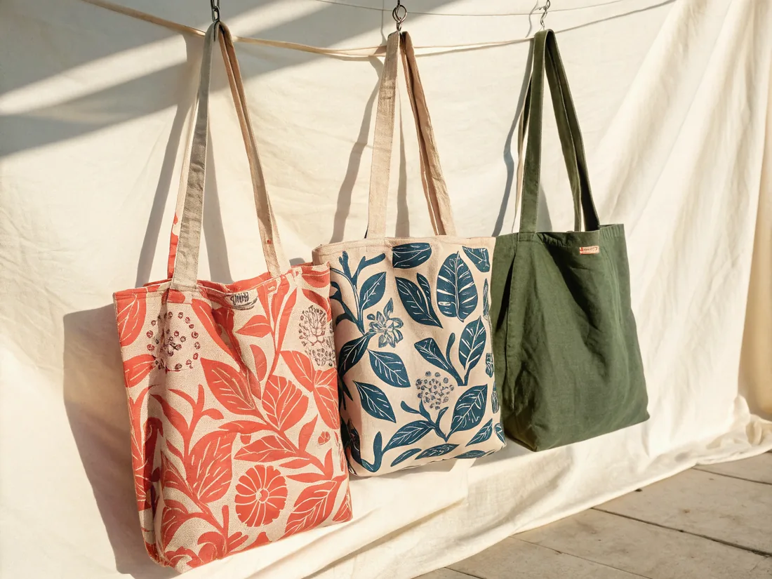 tres bolsas de tela colgadas de un cordel frente a un lienzo blanco al sol, dos estampadas con un follaje a una tinta, coral y azul petróleo, y la tercera de lino verde olivo lisa