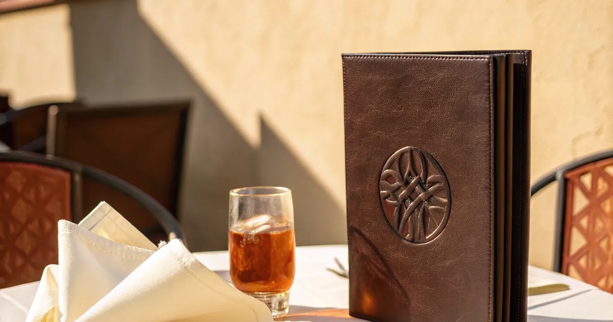 porta carta de piel café de pie sobre una mesa de terraza con mantel blanco, con un emblema circular de trazo entrelazado hundido en seco al centro de la tapa y costura fina en todo el borde