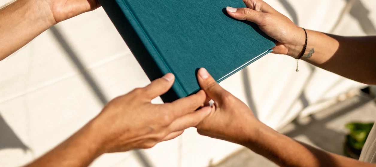 dos pares de manos sostienen un libro de tapa dura forrado en tela verde petróleo bajo sol directo, con las sombras de una celosía sobre el muro claro del fondo