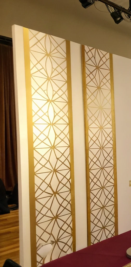 muro de paneles blancos de doble altura con una celosía dorada de trazos entrelazados y bandas verticales doradas entre panel y panel, montado como fondo de un salón con una cortina oscura al costado