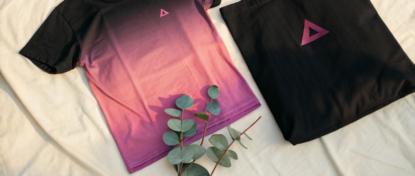 playera negra extendida sobre una sábana crema, con un degradado sublimado que pasa de negro en los hombros a magenta y coral en la bastilla, y un pequeño triángulo magenta en el pecho