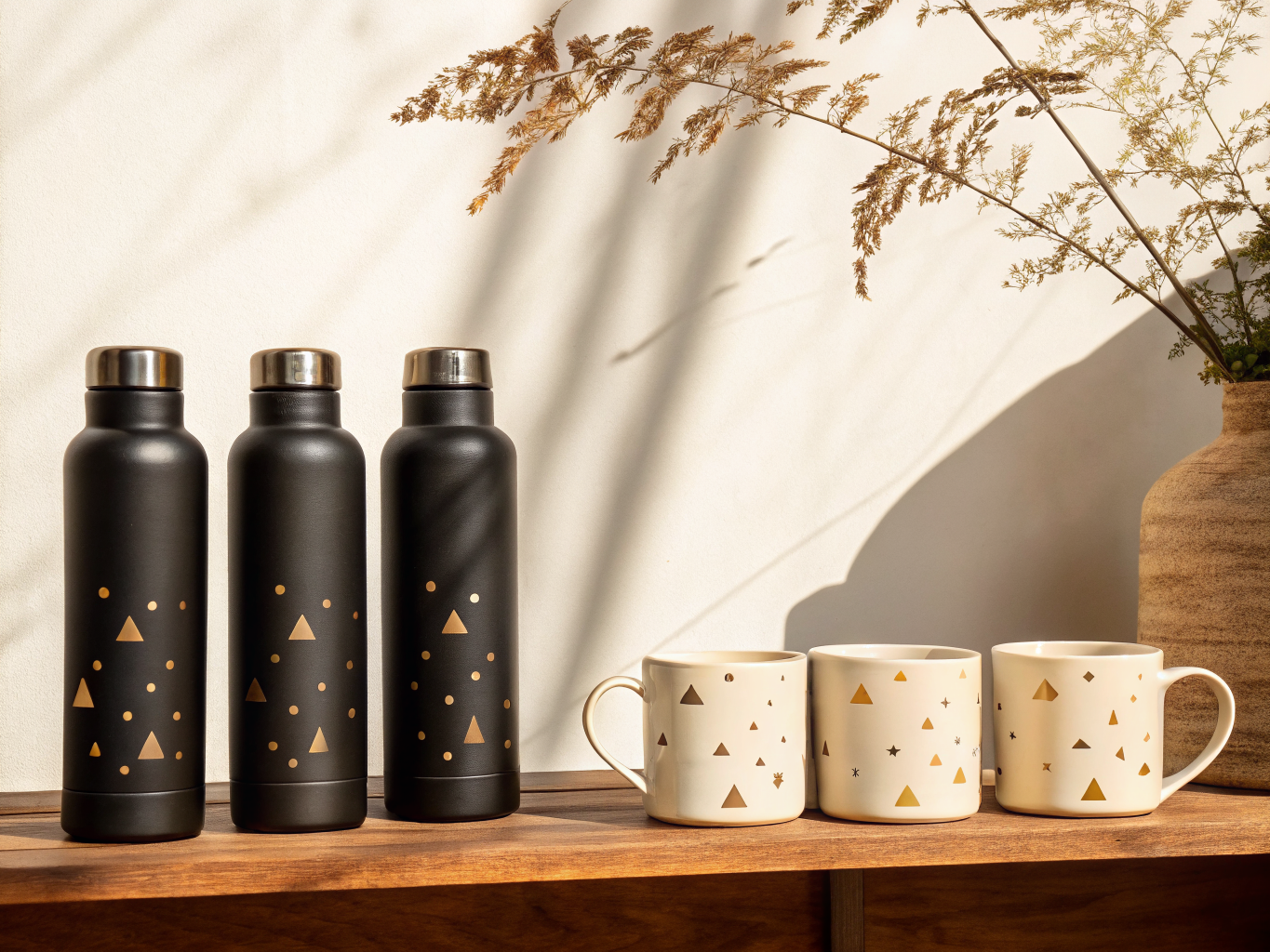 dos termos de acero negro mate y dos tazas de cerámica crema sobre una repisa de madera, todos con un confeti de triángulos y puntos dorados aplicado, con una rama seca proyectando su sombra en el muro