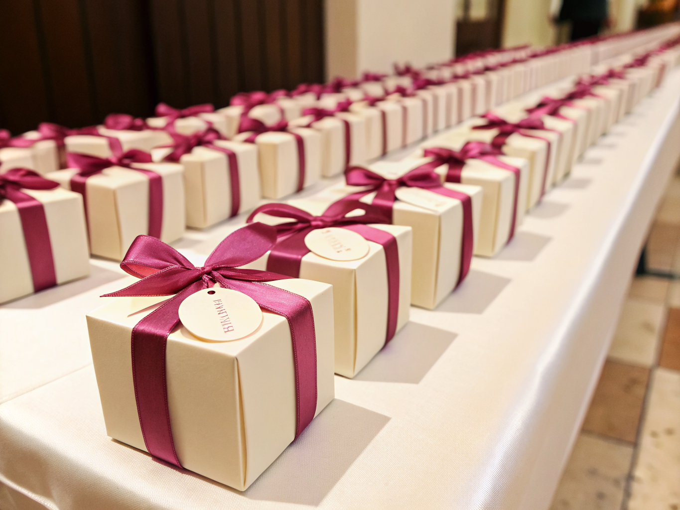 hilera larga de cajas rígidas color crema con listón magenta cruzado y una etiqueta redonda colgando del moño, alineadas sobre una mesa vestida de blanco