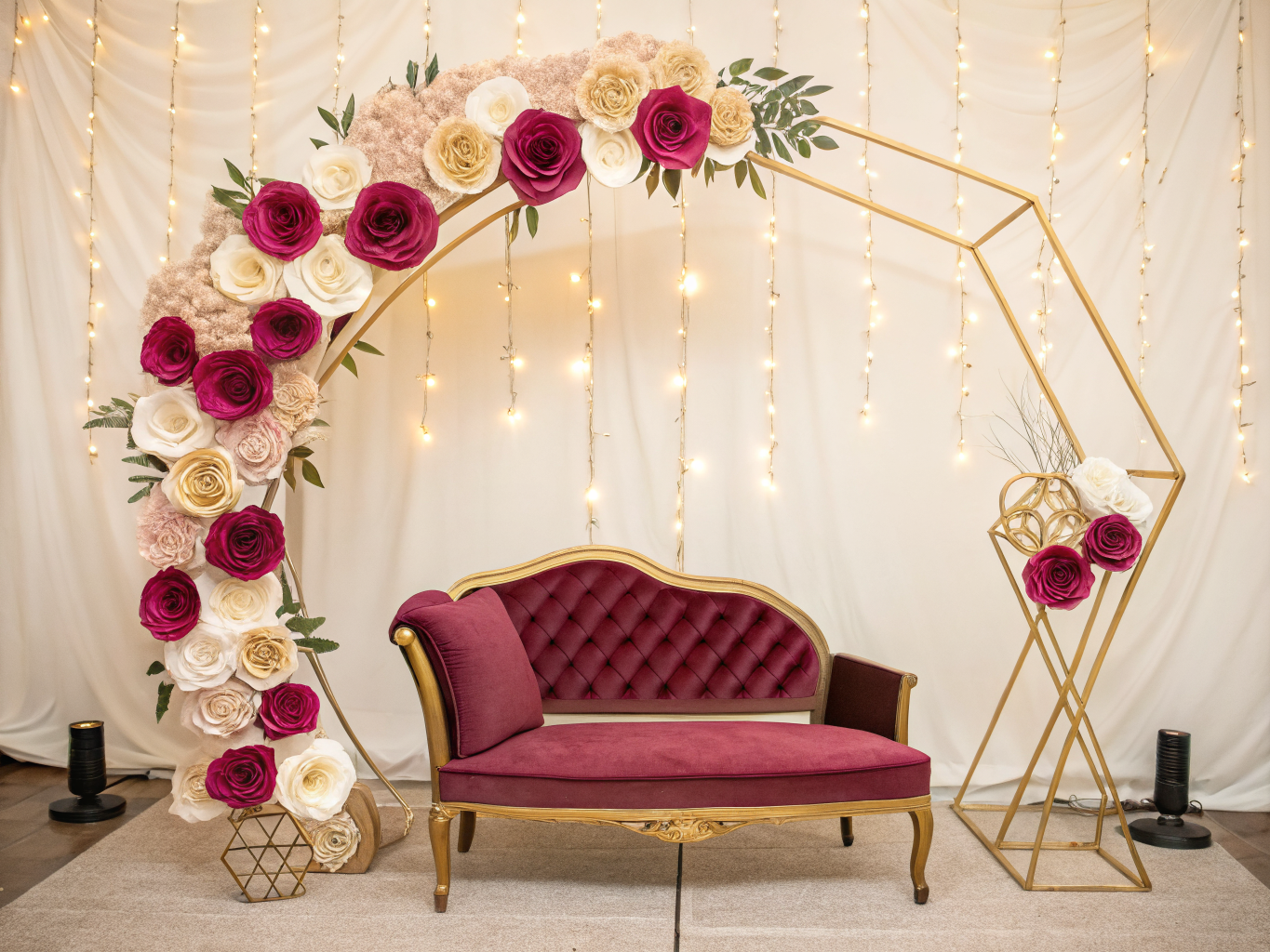 Arco de metal dorado cubierto de rosas gigantes de papel cortadas a mano, con un sofá capitonado vino al centro como fondo de fotos.