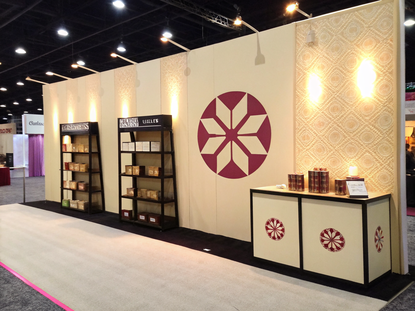 muro de stand de feria en color crema con un emblema geométrico de seis puntas en vino a gran escala, un panel de textura iluminado a un costado y un mostrador con rosetones impresos