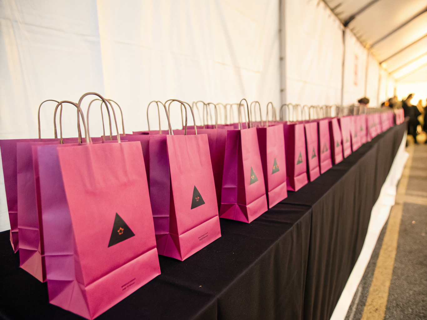 hilera larga de bolsas de papel en magenta y morado con asas de papel torcido y un emblema triangular negro, alineadas sobre una mesa vestida de negro dentro de una carpa