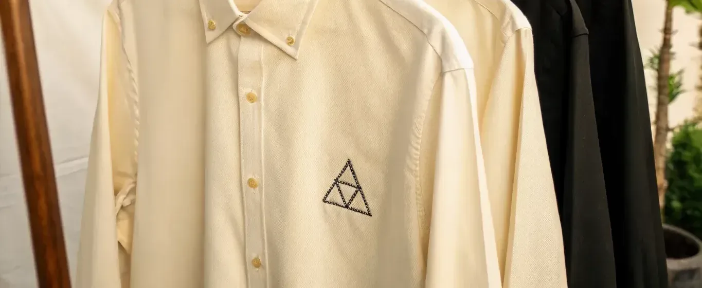 camisa de oxford color crudo colgada en un perchero, con el cuello button down abotonado, la tapeta recta con sus botones y la manga cayendo a la izquierda