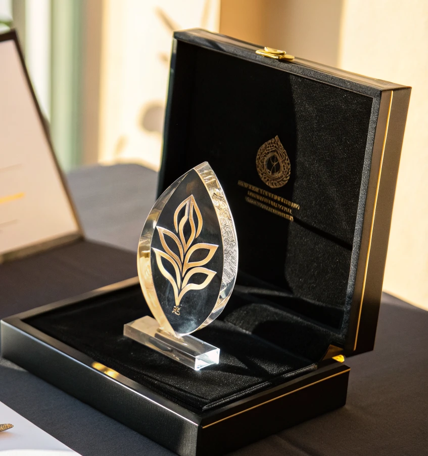 reconocimiento de cristal en forma de hoja con un emblema vegetal grabado y dorado en el centro, montado en su base y presentado dentro de un estuche negro lacado con interior de terciopelo y un emblema dorado en la tapa