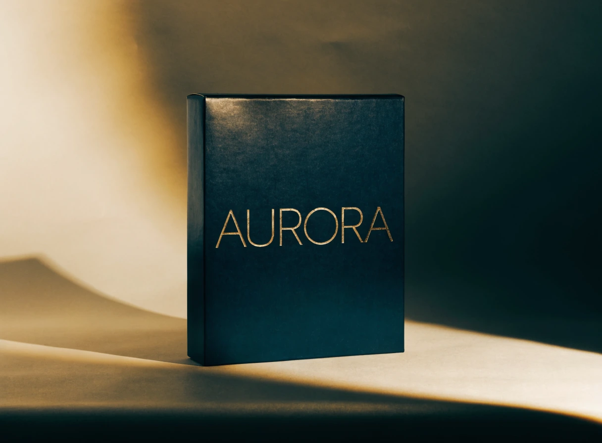 caja rígida azul marino de pie sobre una mesa clara, con el nombre AURORA en foil dorado hundido en el papel teñido, bajo una luz rasante que marca el hueco de cada letra