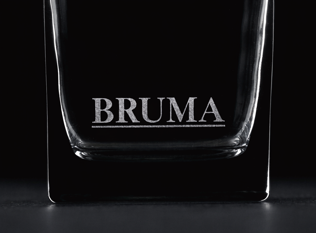 Base de un vaso cuadrado de cristal grueso fotografiada de cerca sobre fondo negro, con el nombre BRUMA arenado en positivo y una línea fina debajo del texto.