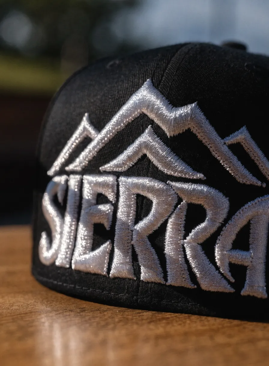 gorra negra con el nombre Sierra y un logo de montañas bordado en 3D con hilo plateado, en acercamiento sobre una mesa de madera