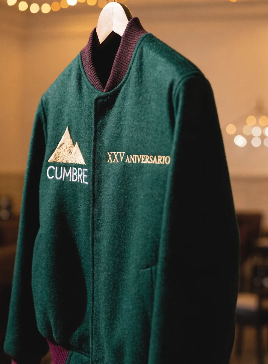 chamarra de lana verde en su gancho, con puños y cuello vino, la montaña de la marca Cumbre bordada en hilo dorado y crema junto a XXV Aniversario