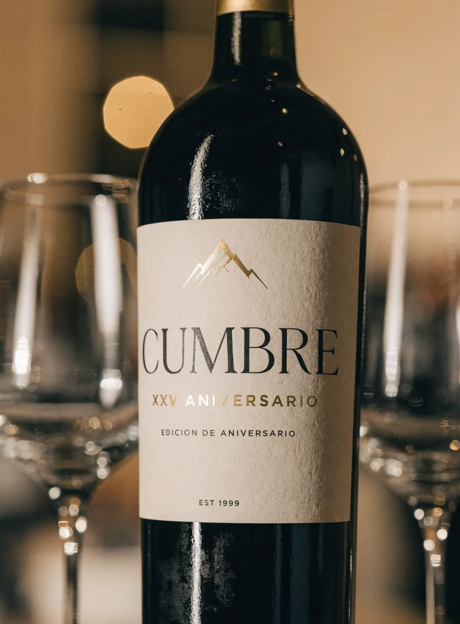 botella de vino tinto con etiqueta crema de la edición de aniversario Cumbre XXV, con la montaña en foil oro, entre dos copas de vidrio