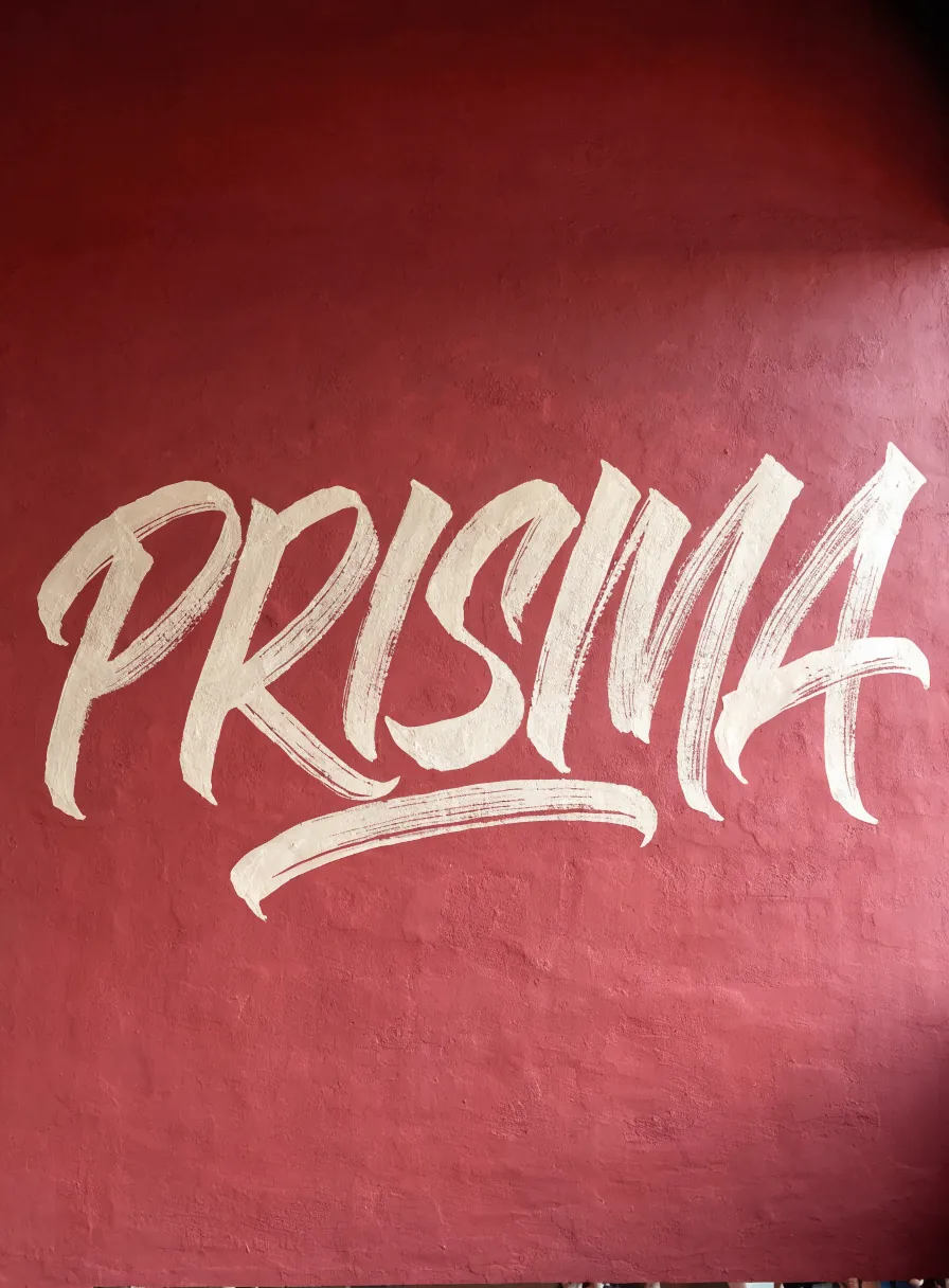 caligrafía a mano con el nombre de la marca ficticia PRISMA en trazo de pincel blanco sobre un muro de estuco terracota