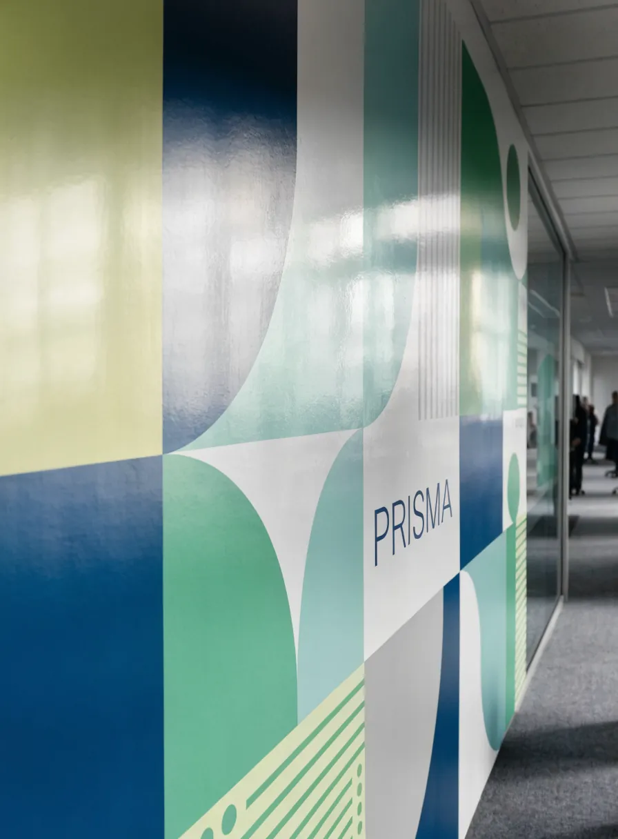 gráfica ambiental de patrón geométrico en azul, verde y lima con el nombre de la marca ficticia PRISMA, aplicada a lo largo del muro de un pasillo de oficinas