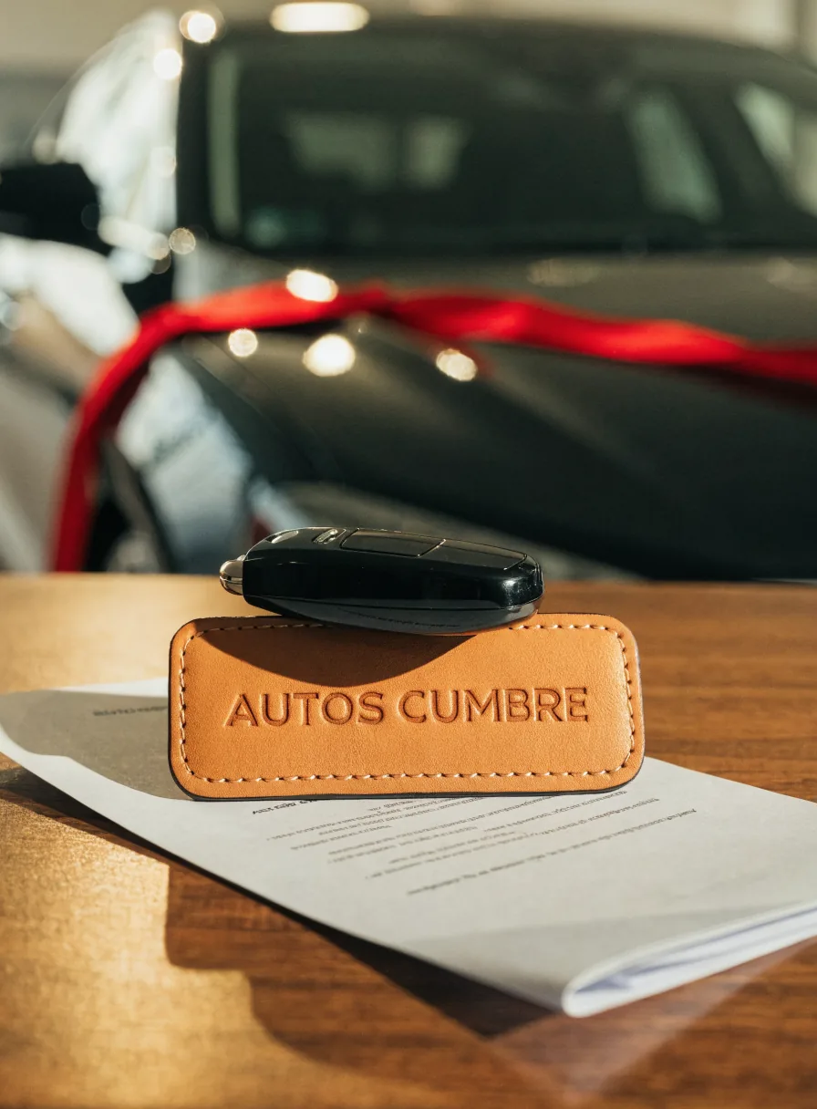 llave del auto sobre un llavero de piel natural repujado Autos Cumbre, apoyado en los documentos de entrega frente a un auto con moño rojo