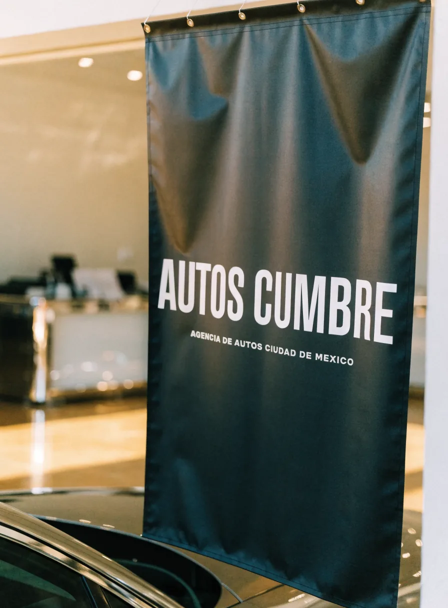 lona colgante de gran formato Autos Cumbre en el piso de ventas, impresa con la marca y la leyenda Agencia de Autos Ciudad de México sobre un auto nuevo