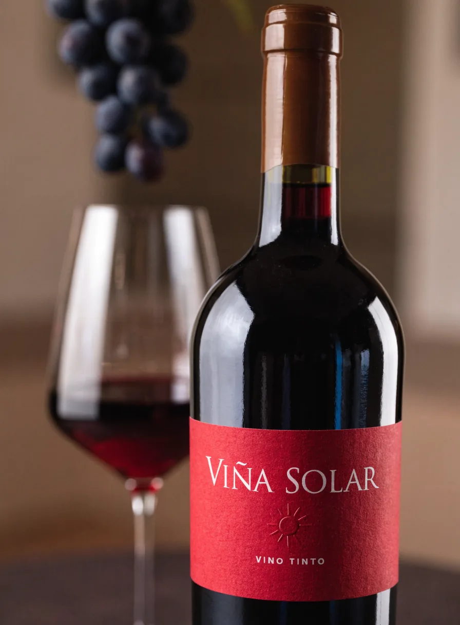 botella de vino tinto con etiqueta roja Viña Solar, Vino Tinto, junto a una copa servida y un racimo de uvas
