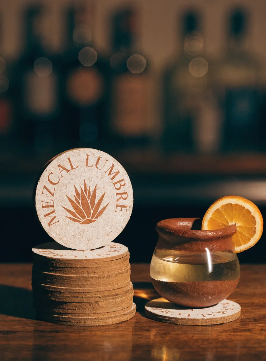 pila de posavasos de pulpa prensada impresos Mezcal Lumbre, junto a una copita de barro con mezcal y una rodaja de naranja