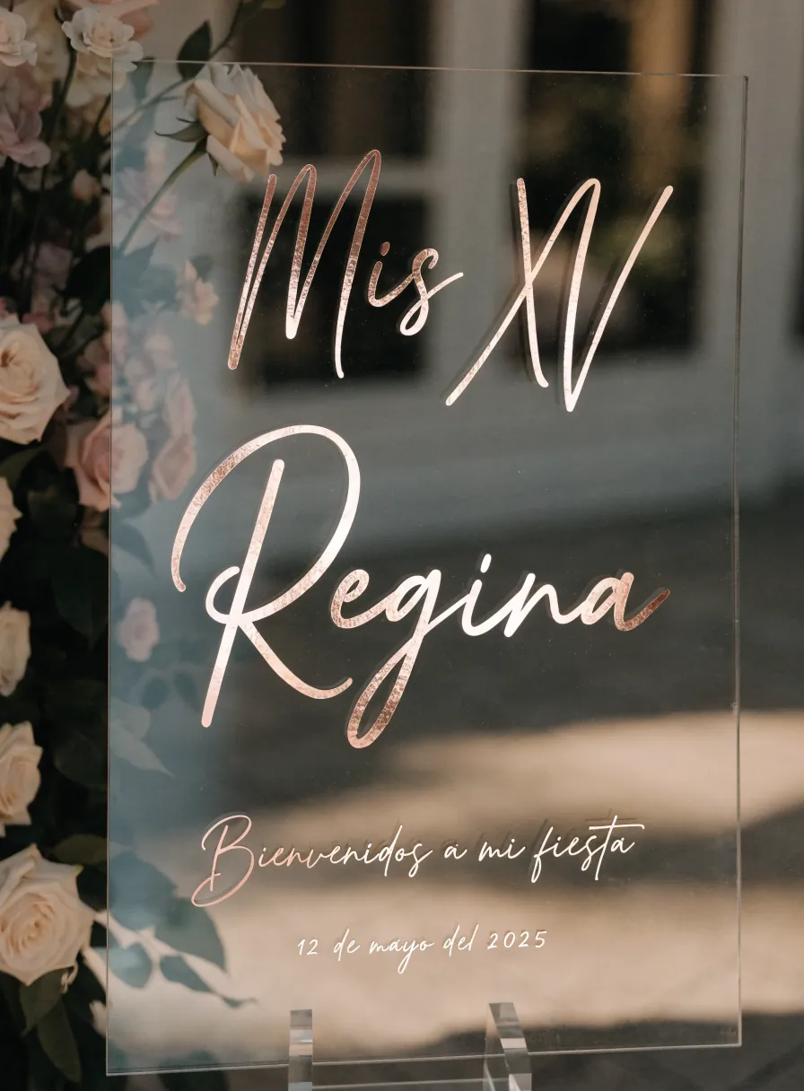 letrero de bienvenida en acrílico transparente con Mis XV Regina en foil color oro rosa, bienvenidos a mi fiesta y la fecha, entre rosas