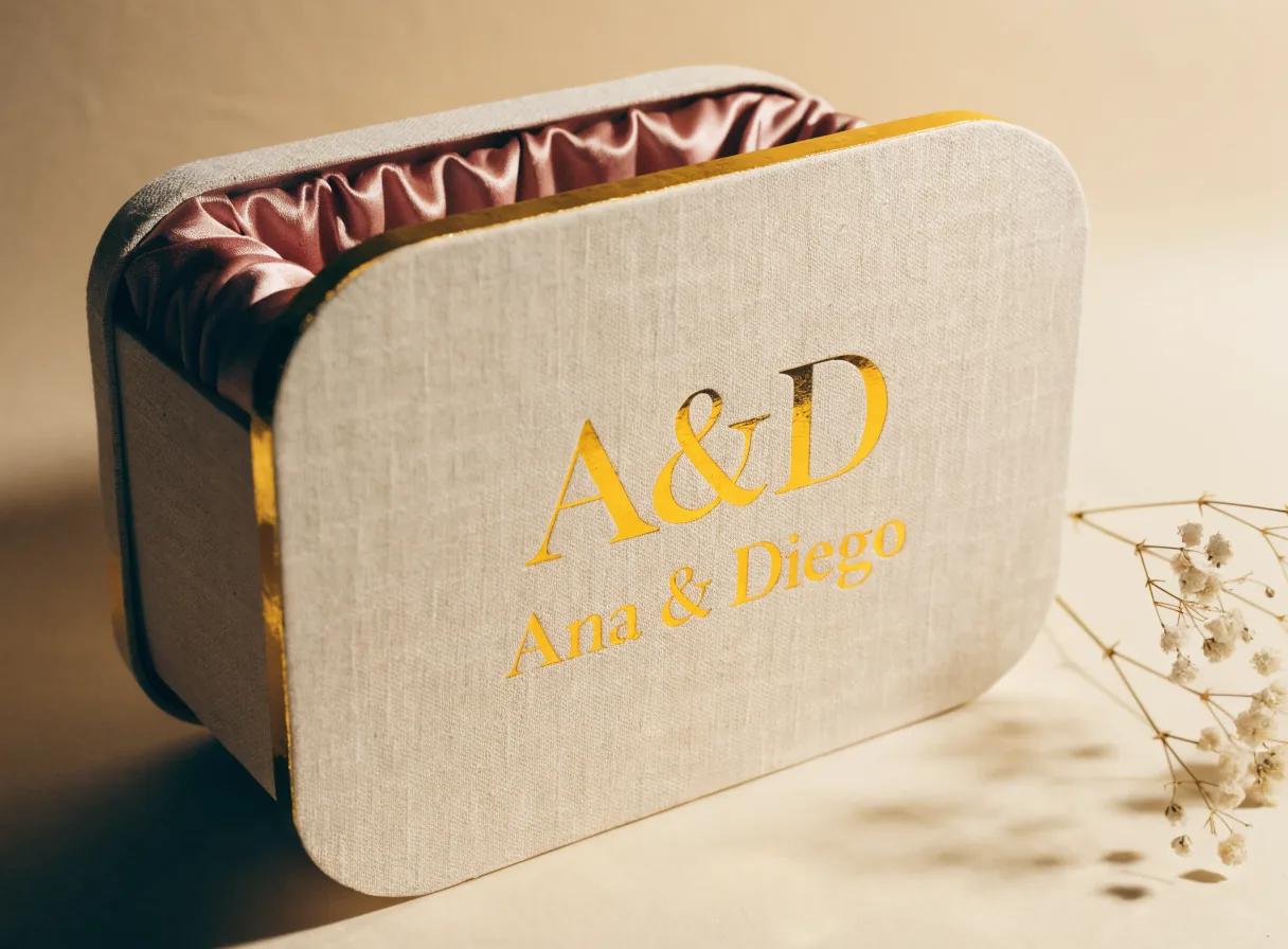 caja rígida de lujo en lino color hueso con el monograma A&D y el nombre Ana & Diego en foil dorado, interior de satín rosa y una ramita de nube