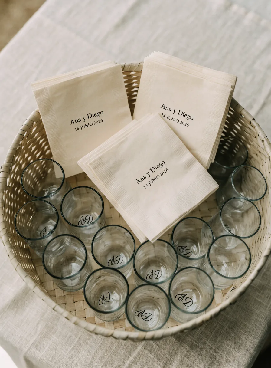 canasta llena de servilletas de coctel y vasitos de cristal, todos impresos con el nombre Ana y Diego y la fecha, uno para cada invitado