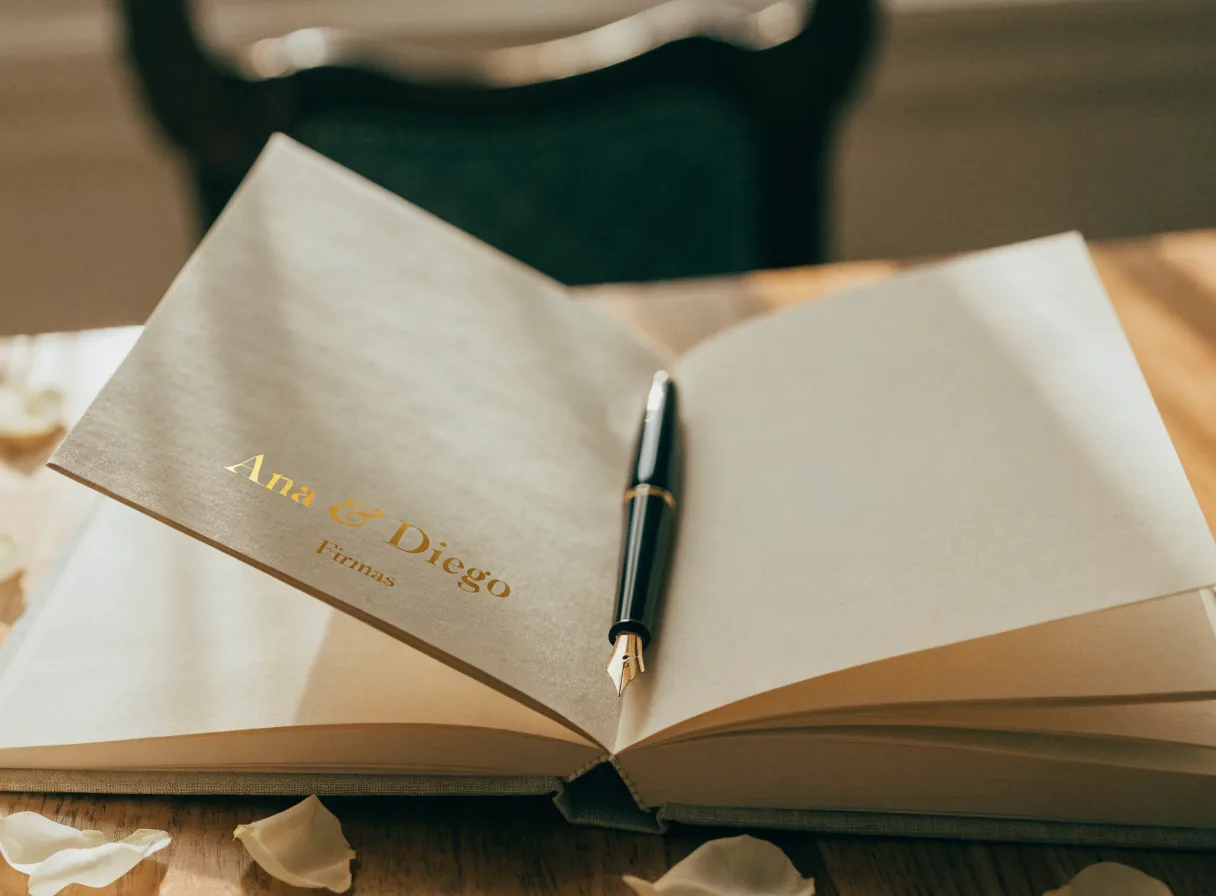libro de firmas abierto en páginas de algodón en blanco, con una pluma fuente descansando y la portada de lino estampada en foil dorado con Ana & Diego y la palabra Firmas, entre pétalos