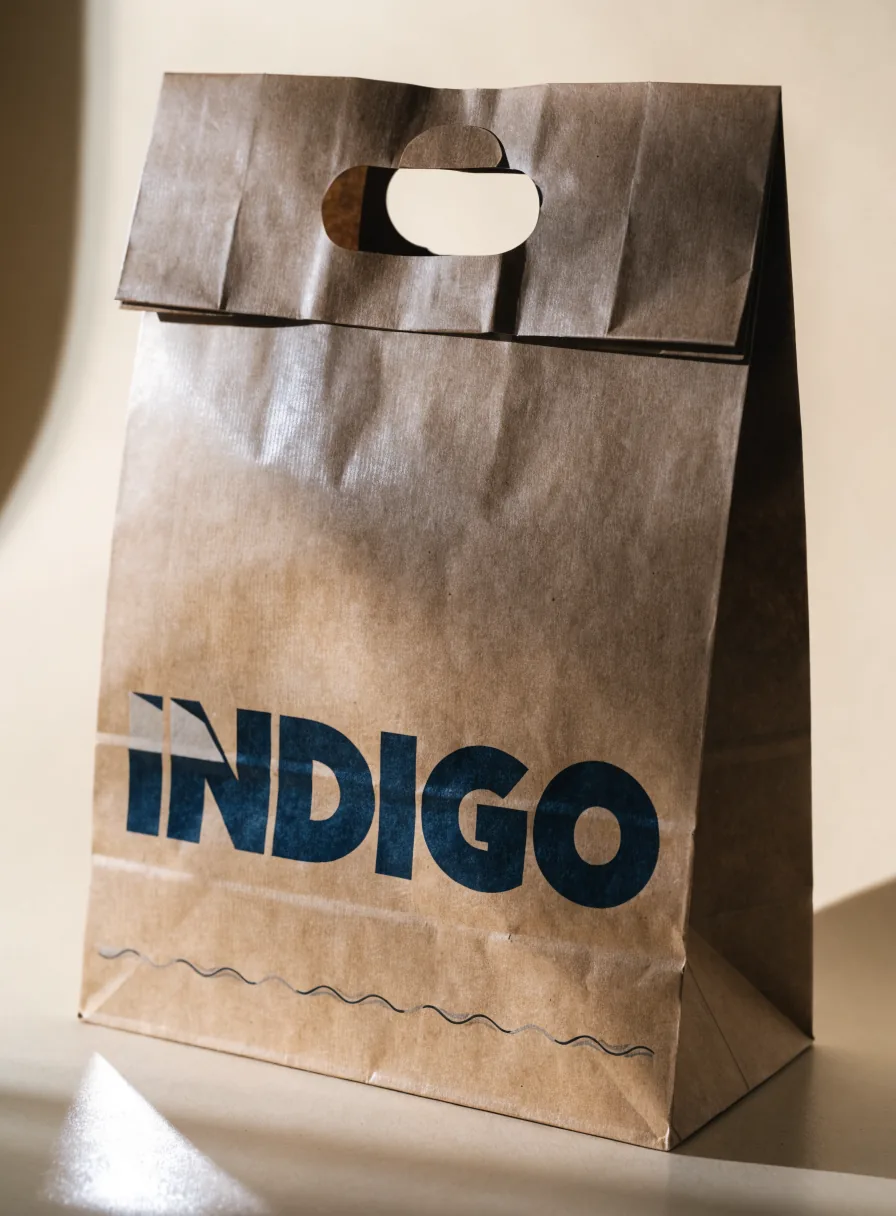 bolsa de papel kraft con asa troquelada ovalada reforzada y la palabra INDIGO impresa en azul marino sobre una onda al pie, sobre fondo crema con luz cálida