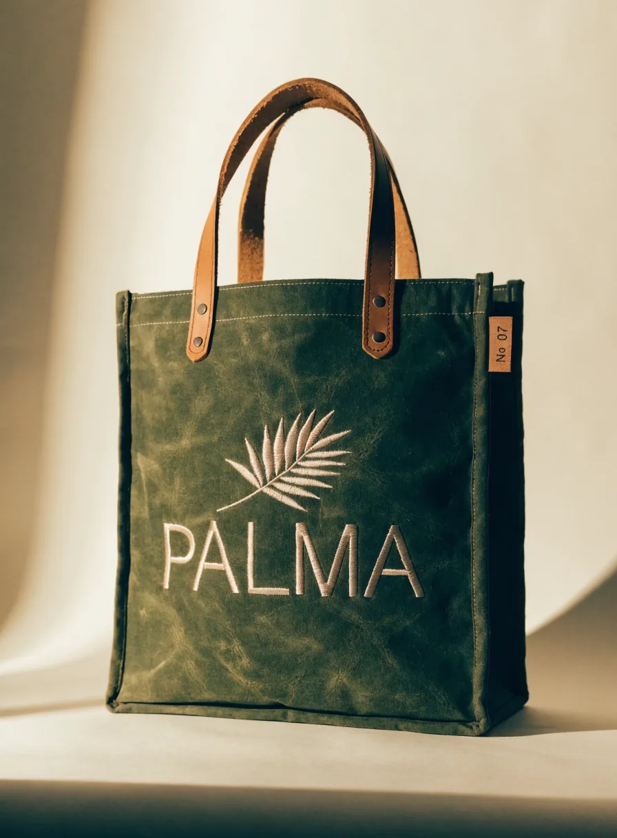 bolsa boutique estructurada de lona encerada verde olivo con asas de piel color caramelo remachadas, una hoja de palma y la palabra PALMA bordadas en hilo crudo, y una etiqueta de piel numerada No 07 cosida al costado, sobre fondo crema con luz cálida