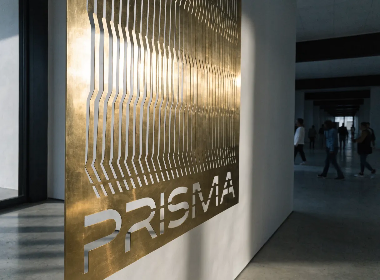 biombo de latón cortado a láser con líneas onduladas de tipo topográfico y la palabra PRISMA calada en la base, en un pasillo de galería