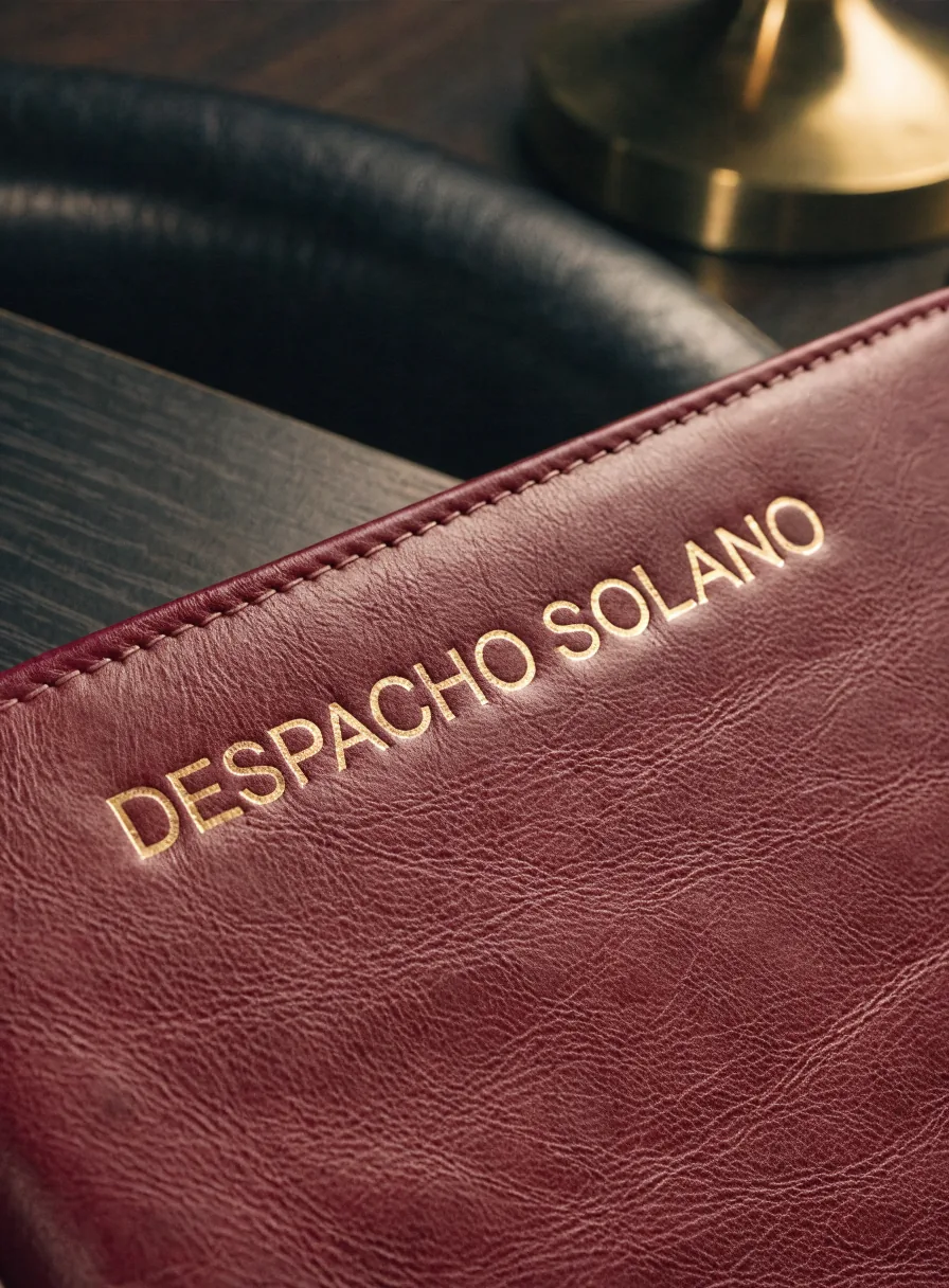 portafolio de piel color vino con el nombre Despacho Solano en foil dorado, sobre un escritorio oscuro junto a una lámpara de latón