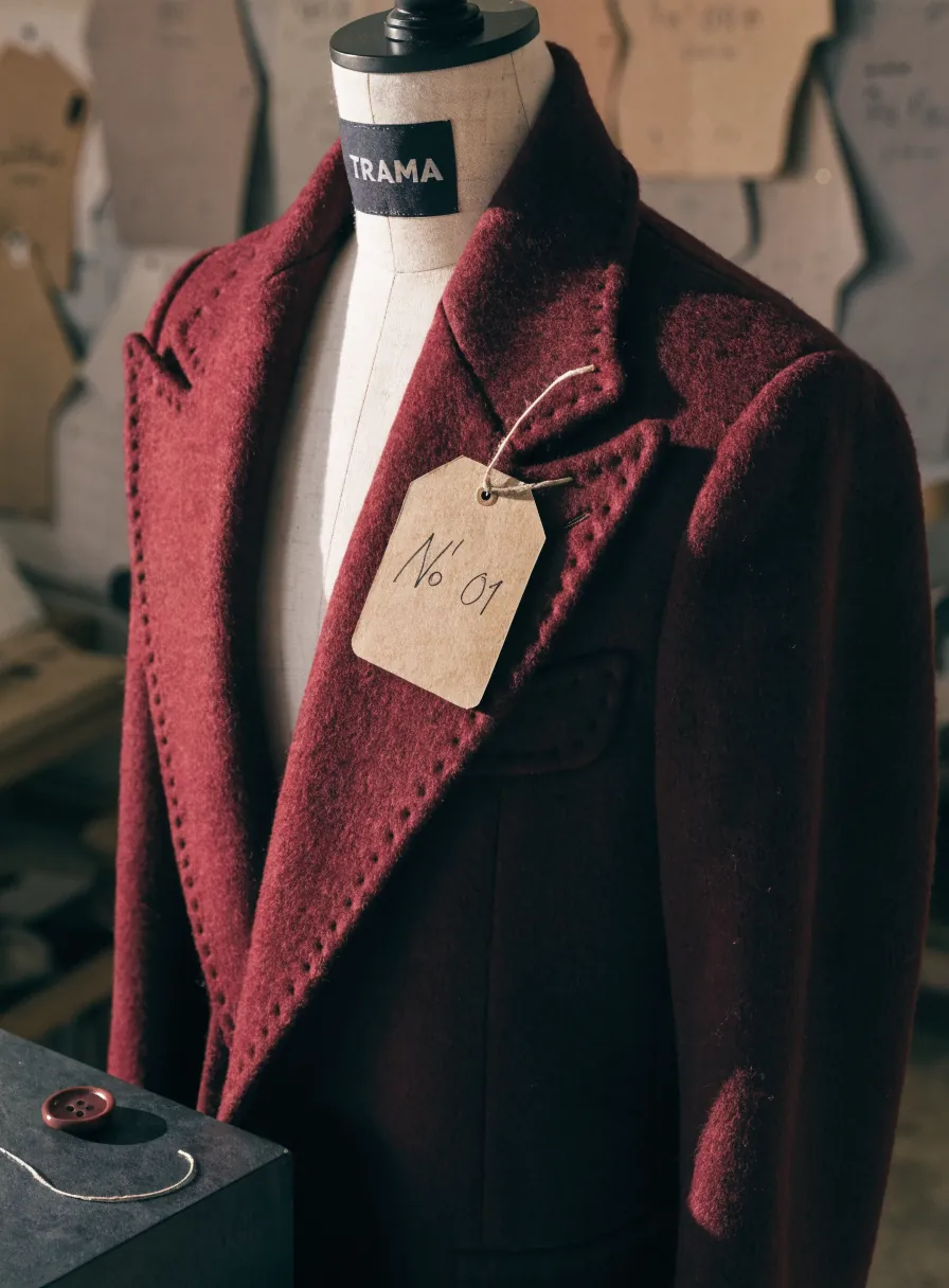 saco de autor en lana color vino sobre un maniquí de sastre con la etiqueta TRAMA, pespunte a mano en las solapas y una etiqueta kraft Nº 01, en un atelier con patrones al fondo