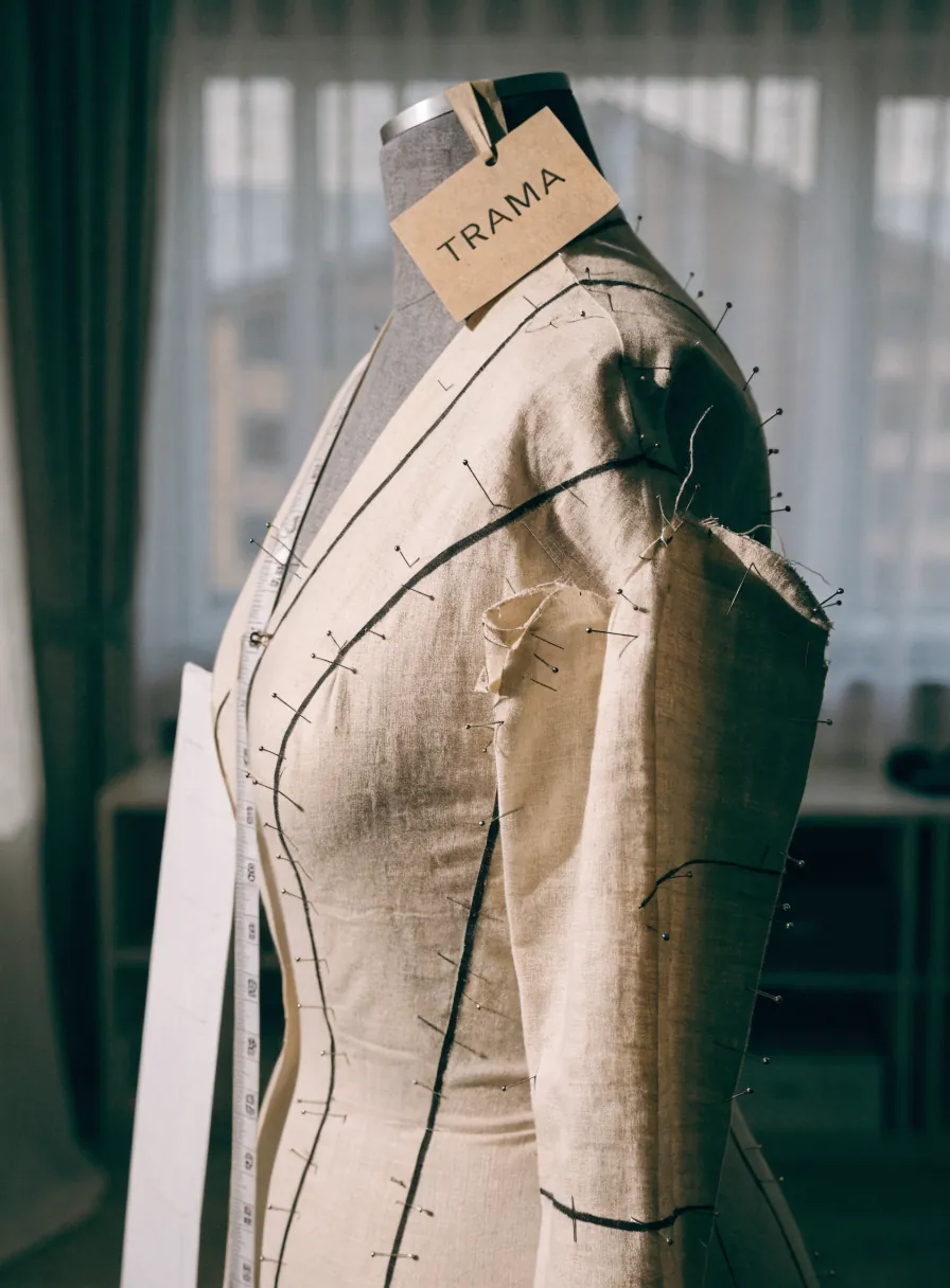 toile de ajuste en lino crudo alfilerado sobre un maniquí de sastre, con cinta métrica, líneas de costura marcadas y una etiqueta TRAMA, en el atelier