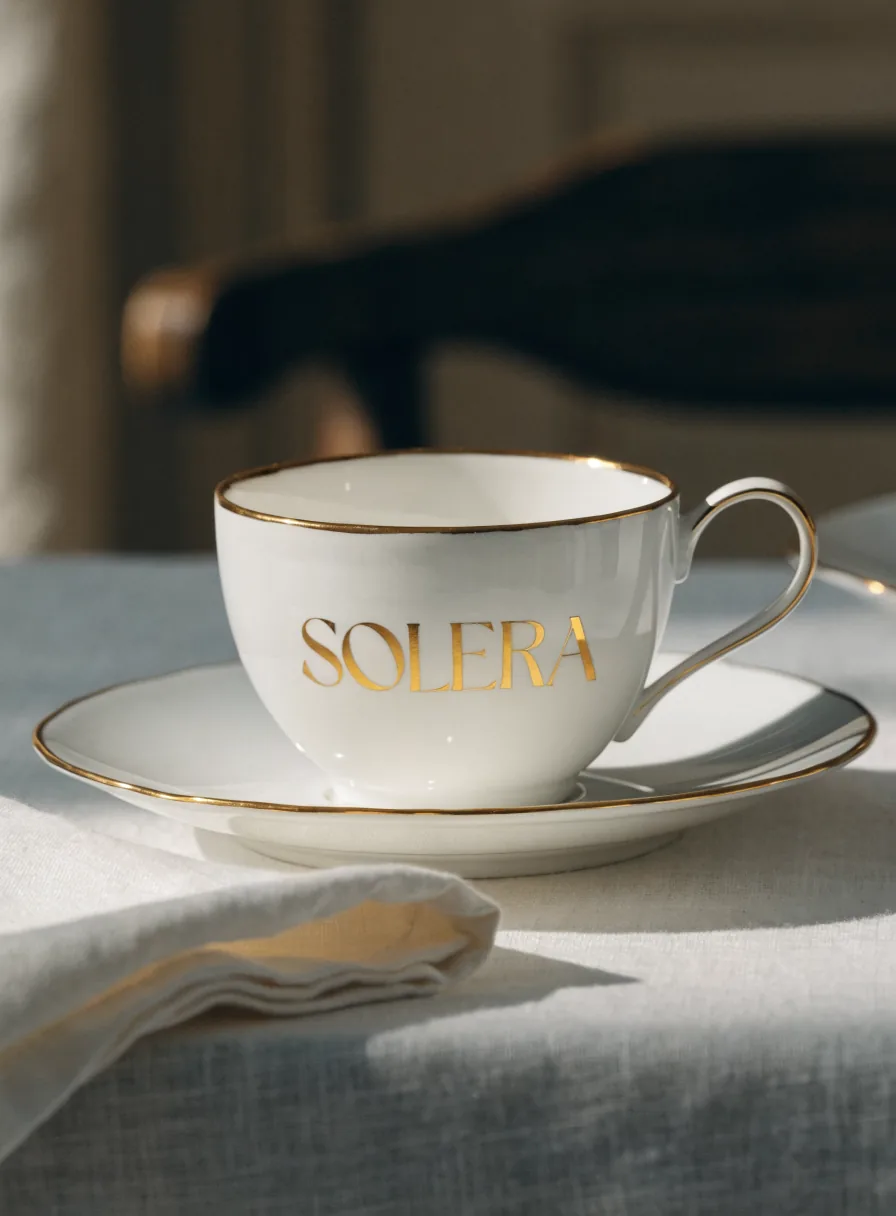 taza y plato de porcelana con filo de oro y la marca ficticia Solera en foil dorado, sobre una mesa vestida de lino