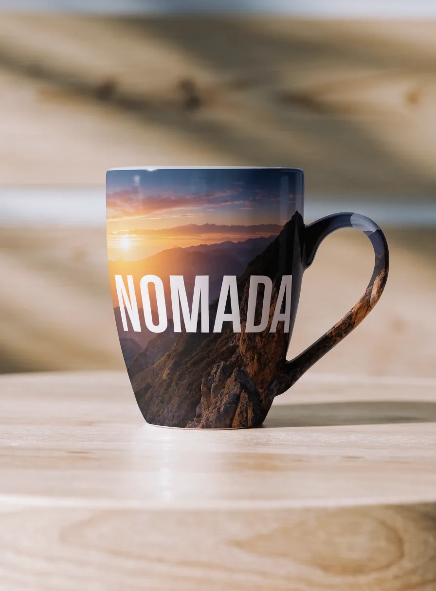 taza de cerámica sublimada de orilla a orilla con la foto de un atardecer entre montañas y la marca NOMADA, sobre una mesa de madera