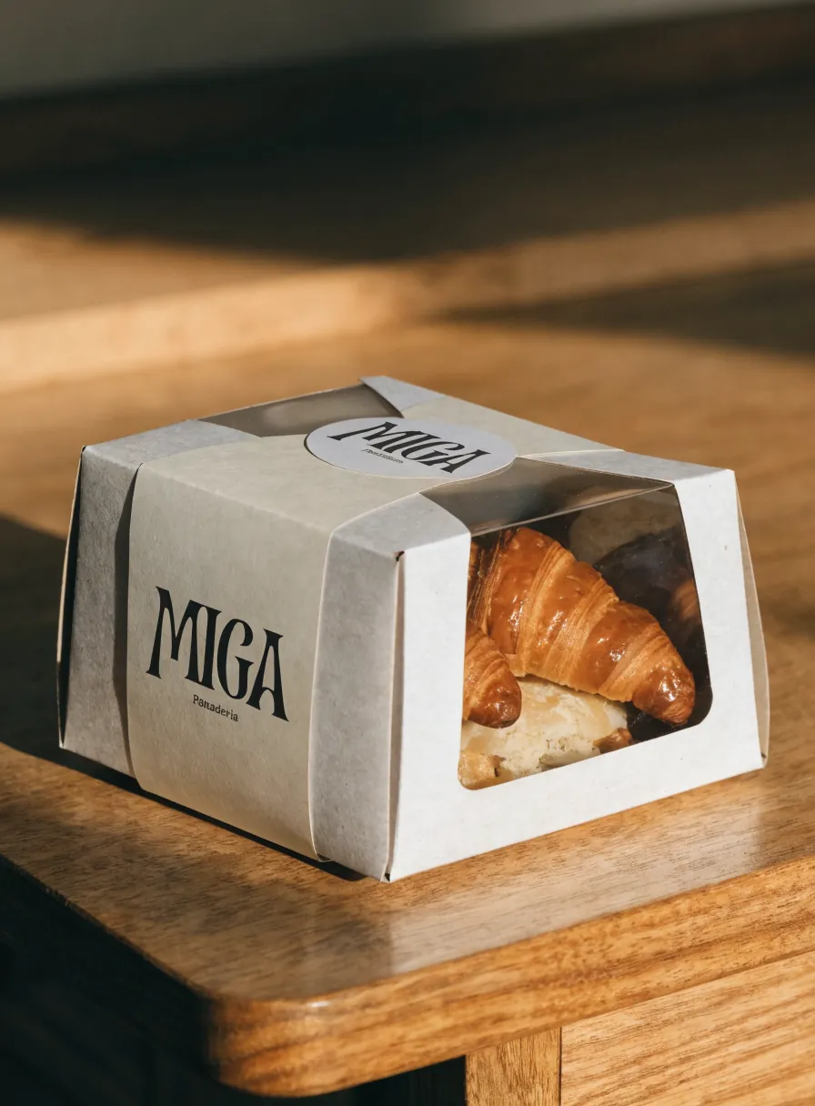 caja de repostería para llevar en cartón claro con ventana, una faja impresa con la marca Miga y un sello redondo, con croissants adentro