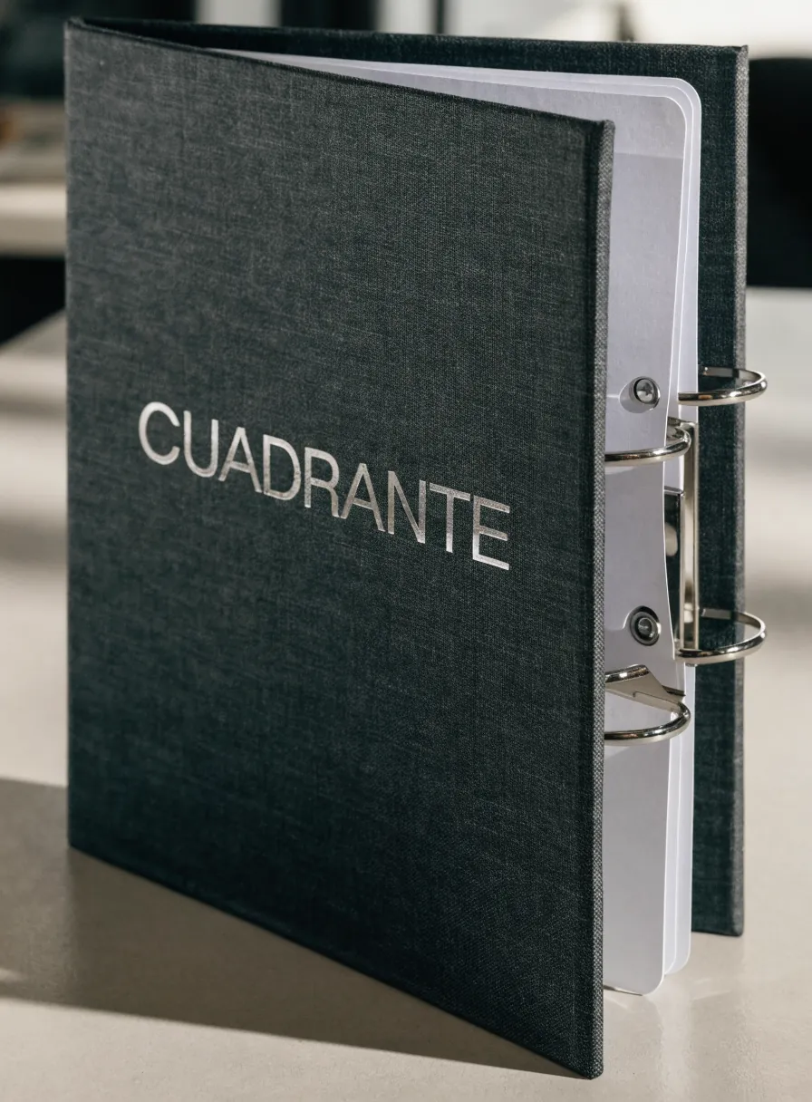 carpeta de argollas forrada en tela gris carbón con el nombre CUADRANTE en foil plata y el mecanismo de tres anillos a la vista