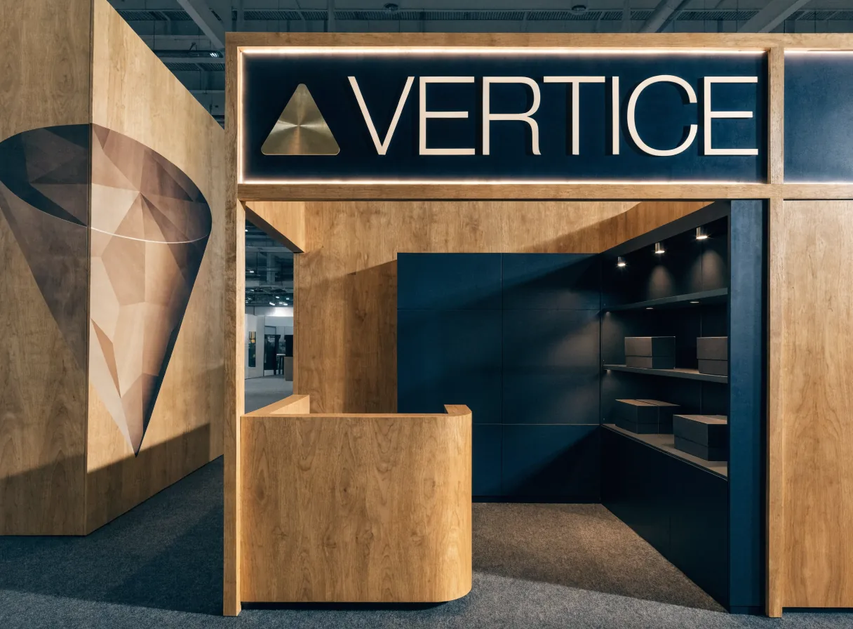 stand de feria construido a la medida con muros de madera, un logotipo Vertice iluminado en el frontón, mostrador de recepción y vitrinas con producto, en un pabellón de expo