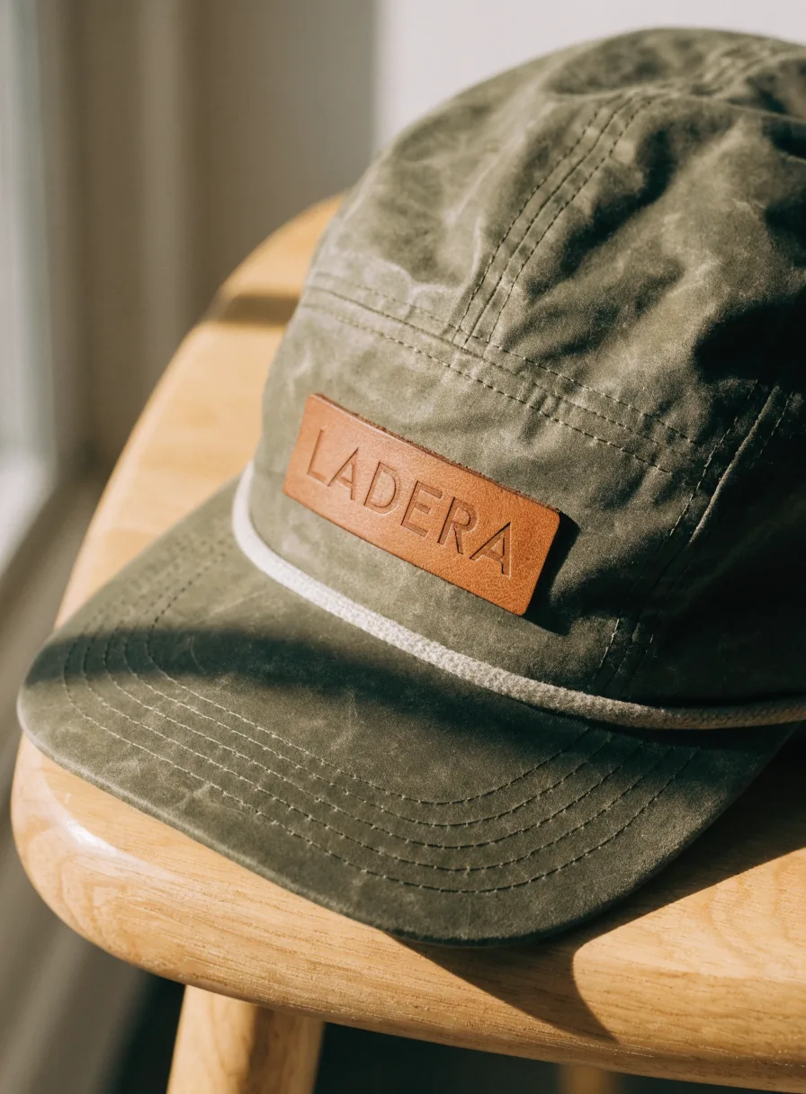 gorra camper de cinco paneles en lona encerada olivo sobre un banco de roble, con un parche rectangular de piel curtida cosido al frente y la marca ficticia LADERA marcada en hueco sobre la piel