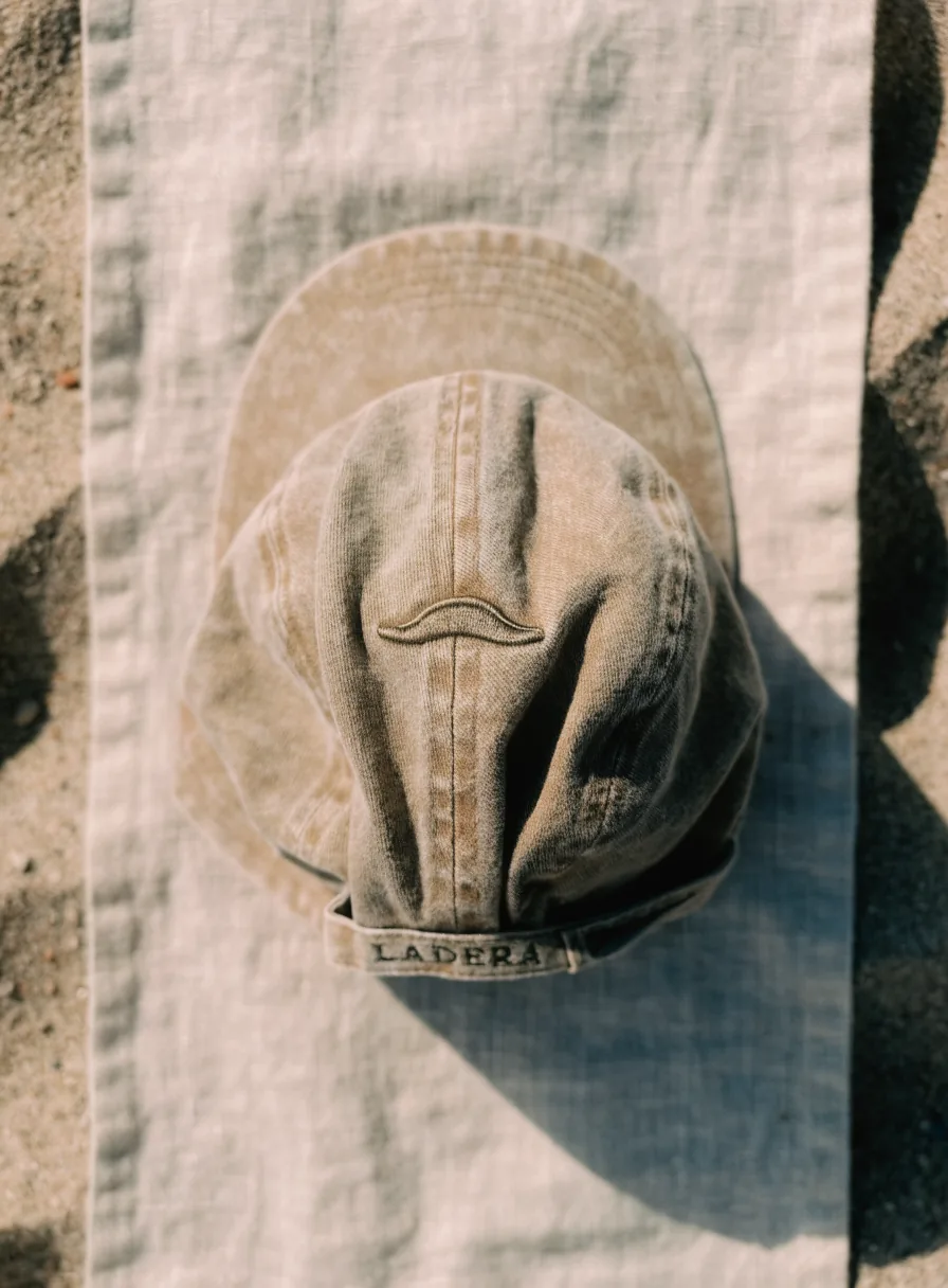 gorra sin estructura de algodón lavado color arena, acostada sobre un lienzo de lino, con la cresta de montaña de la marca ficticia LADERA bordada al frente en hilo olivo y la visera relajada