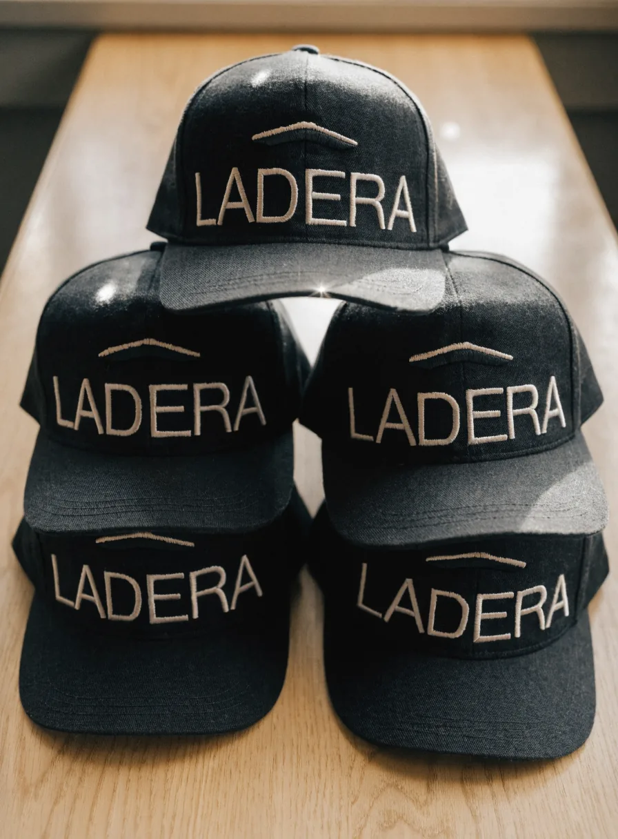 seis gorras estructuradas idénticas en sarga gris carbón apiladas en dos filas sobre una mesa de roble, cada una con la marca ficticia LADERA bordada al frente en hilo crema