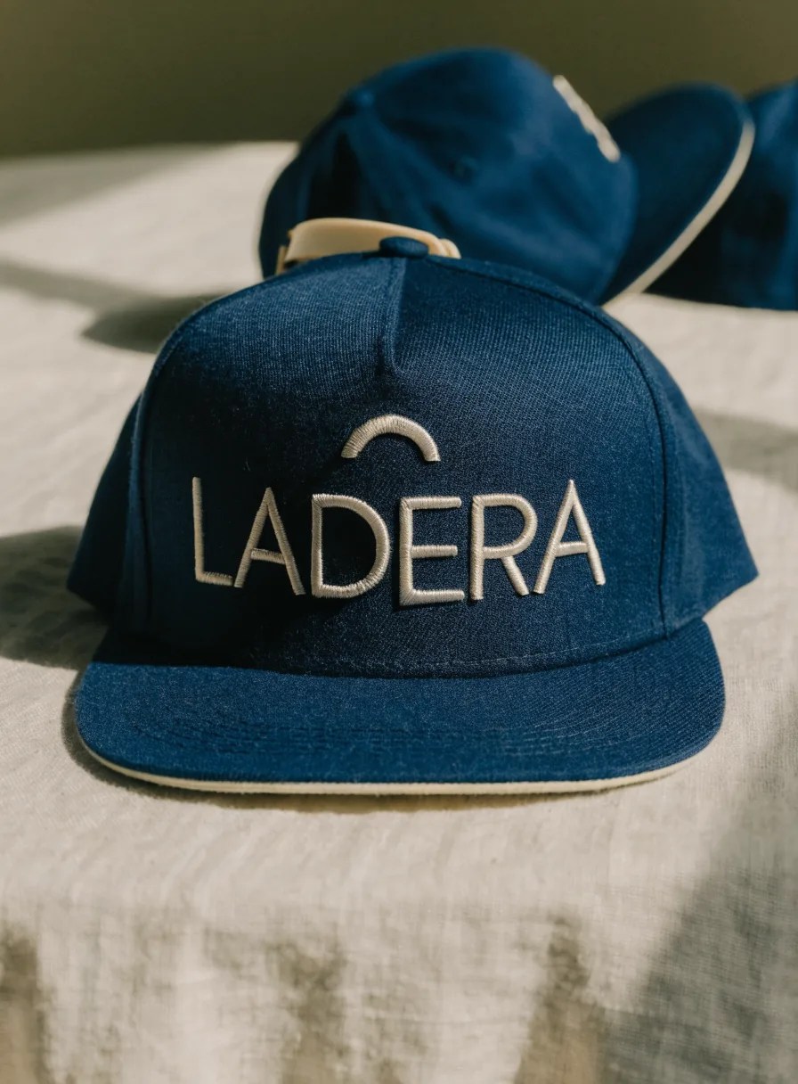 gorra de cinco paneles con visera plana en sarga azul marino de pie sobre lino claro, con la marca ficticia LADERA bordada al frente en punto satín plano con hilo crema, una cresta de montaña bordada arriba y el forro crema bajo la visera a la vista