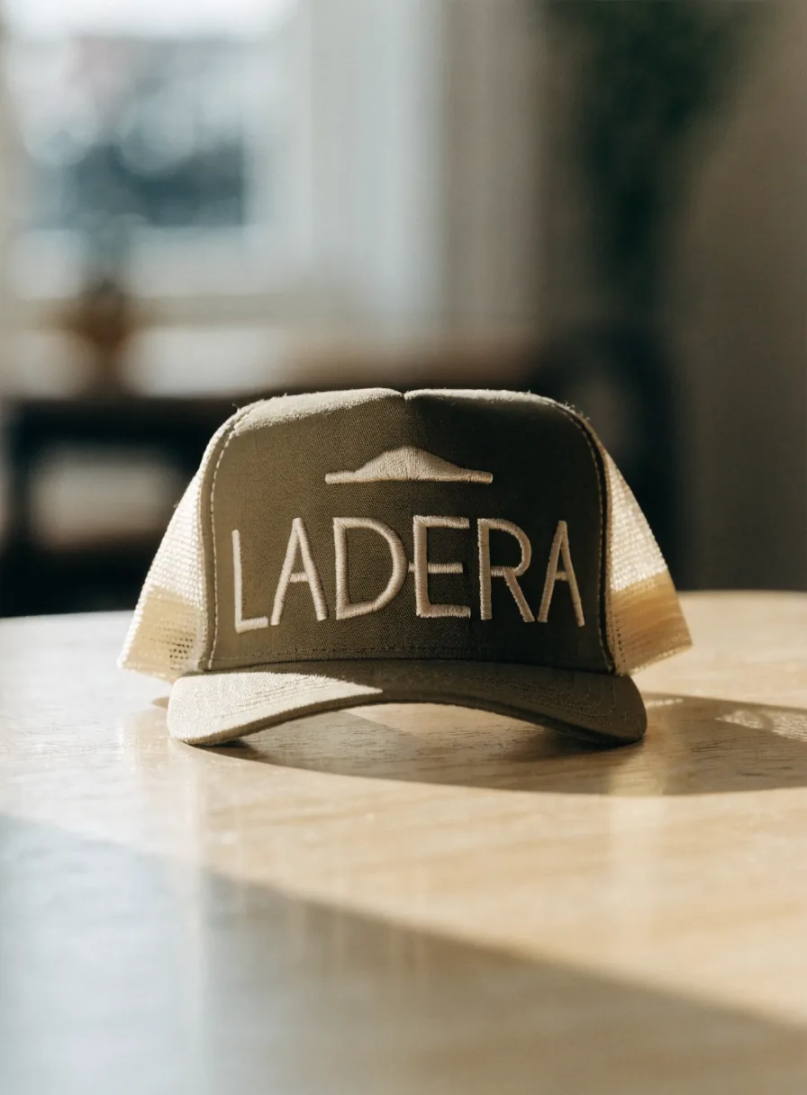 gorra trucker con frente de lona olivo y paneles traseros de malla crema, de pie sobre una mesa de roble, con la marca ficticia LADERA bordada al frente y una cresta de montaña arriba