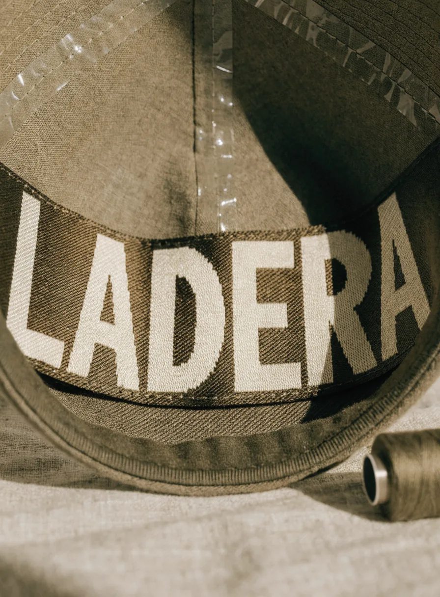 vista cercana del reverso de la visera de una gorra de lona olivo, con la marca ficticia LADERA bordada en grande sobre el forro en hilo crema, y un carrete de hilo del mismo tono al lado