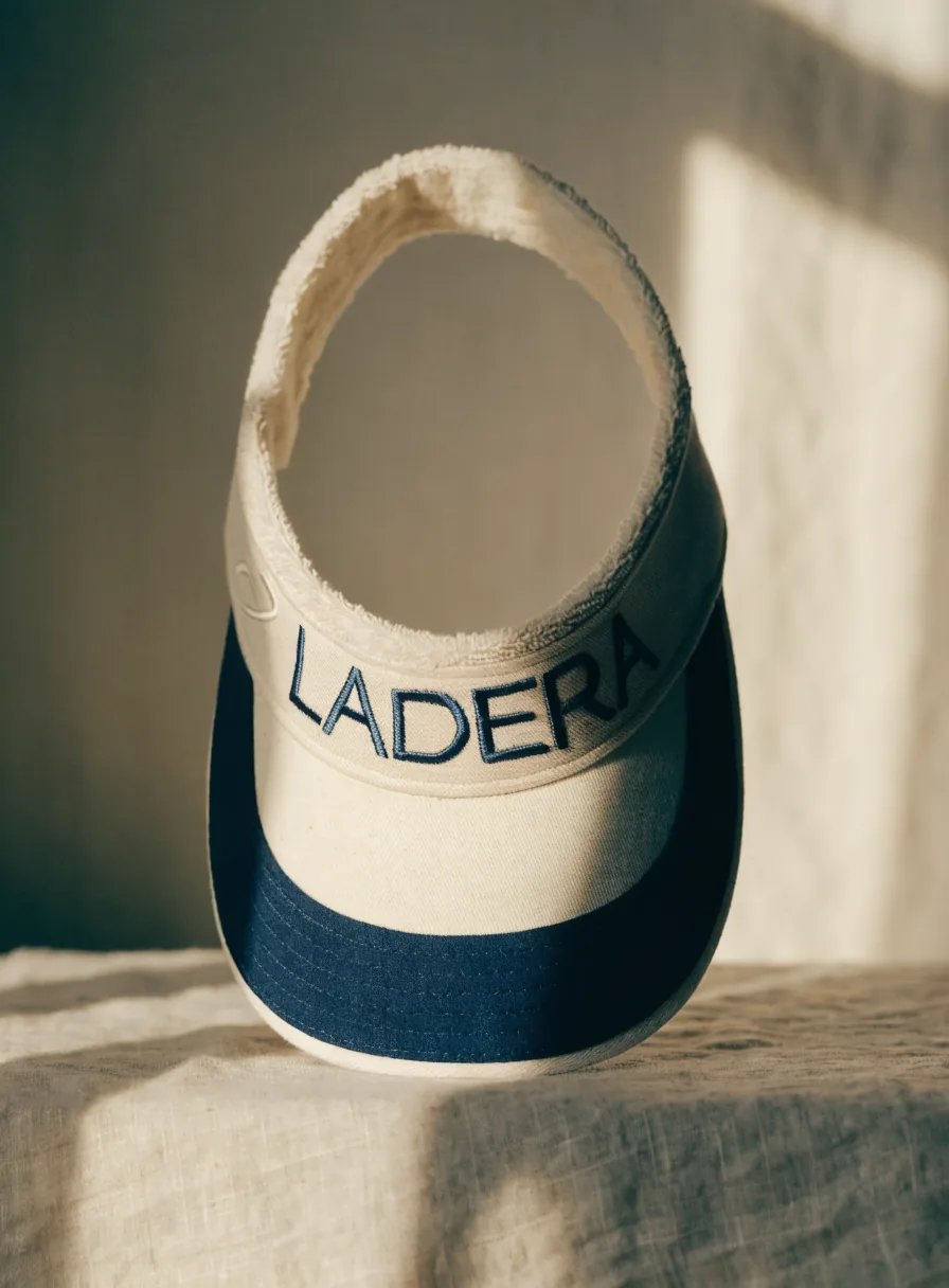 visera de algodón crema con parte inferior azul marino de pie sobre lino, con la banda de toalla a la vista y la marca ficticia LADERA bordada al frente en hilo marino