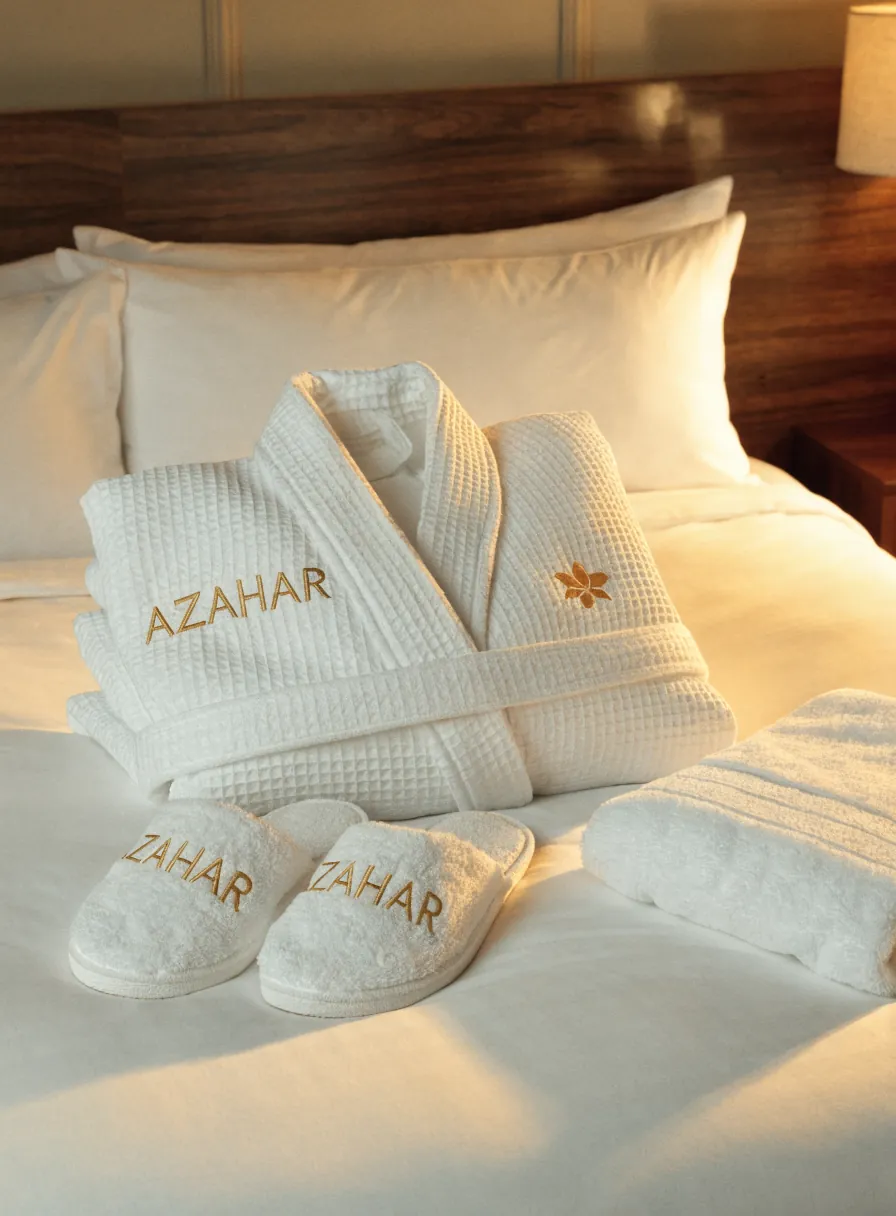 bata de toalla blanca doblada sobre la cama con dos pantuflas bordadas en hilo dorado con la marca Azahar