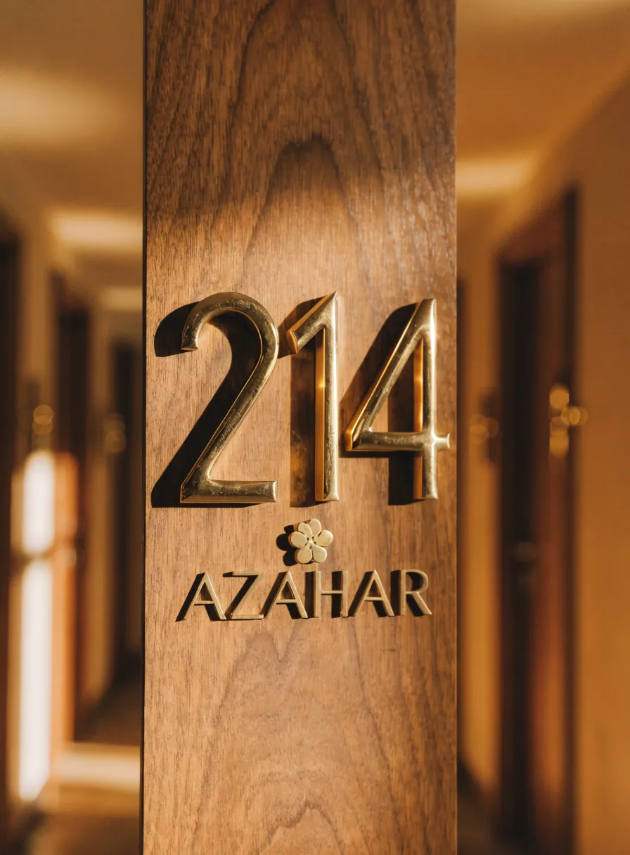 número de habitación 214 en latón dorado y la marca Azahar, montados sobre una puerta de madera de nogal en un pasillo cálido