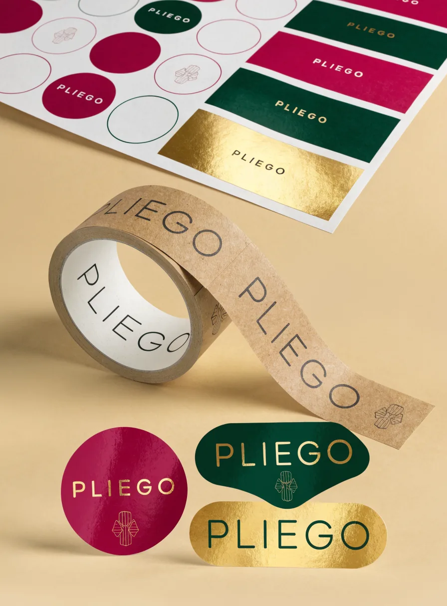 hoja de etiquetas troqueladas de la marca ficticia PLIEGO en magenta, verde y foil dorado, junto a un rollo de cinta de papel kraft impresa y varias calcomanías sueltas