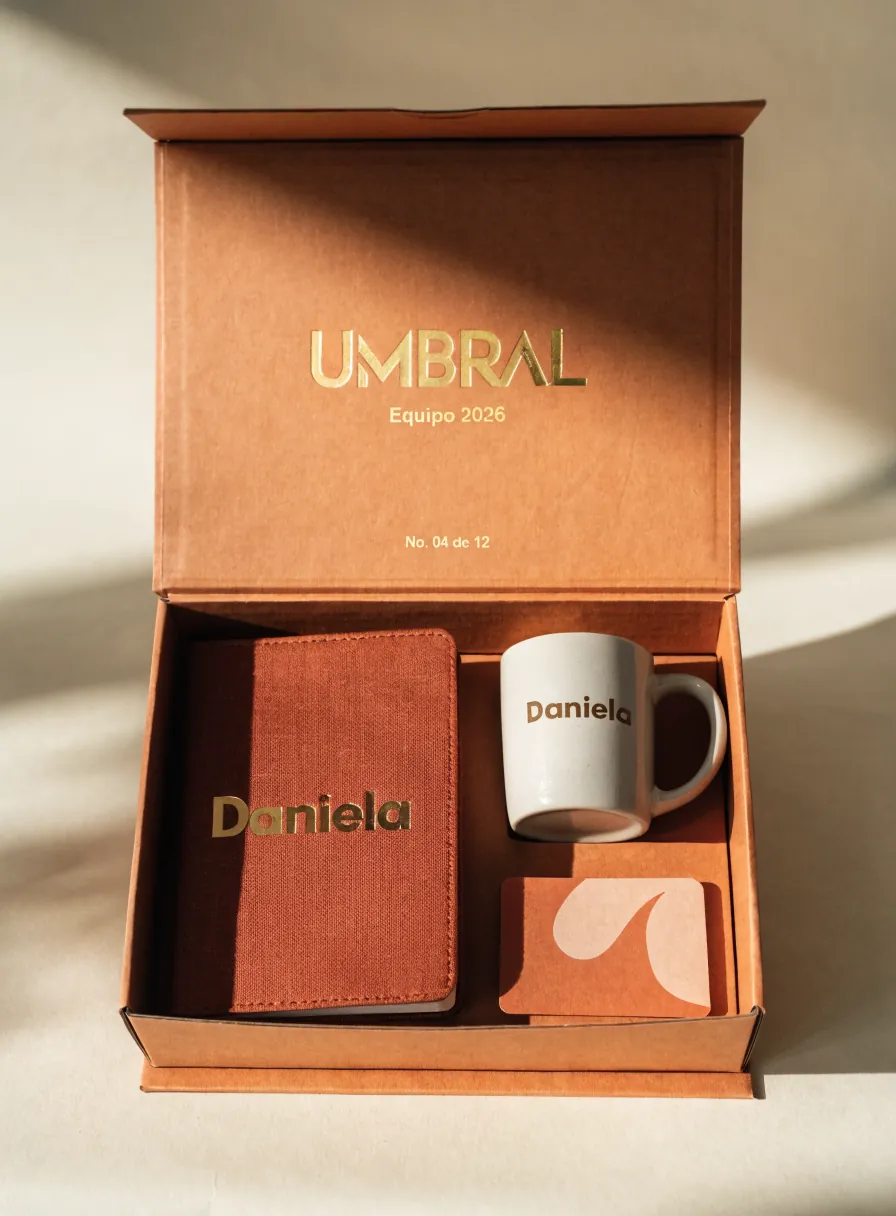 caja de bienvenida abierta con foil dorado UMBRAL, Equipo 2026 y el folio No. 04 de 12, con un cuaderno y una taza grabados con el nombre Daniela dentro