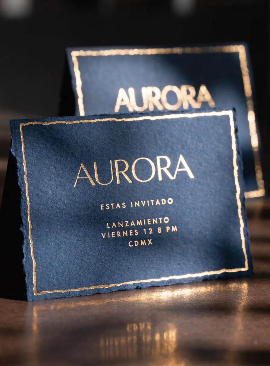 invitación de lanzamiento en papel algodón azul marino de canto deshilachado, con la marca Aurora, la fecha y el lugar en foil dorado